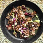 A black frying pan with cooked bacon pieces and green and purple vegetables scattered throughout. The pan is on a textured surface.