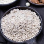 A bowl filled with ground almond meal, placed on a dark surface, with a small portion of whole almonds visible in the background.