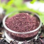 A small glass bowl filled with purple hibiscus powder, surrounded by dried hibiscus petals and green leaves in the background.