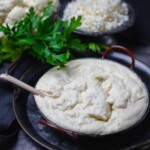 A bowl of creamy cauliflower mash is placed next to fresh parsley and a bowl of crumbled cauliflower.