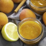 A jar of peach jam next to fresh peaches, a halved lemon, and a cinnamon stick on a gray cloth background.