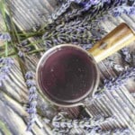 A glass cup containing lavender cocktail syrup, surrounded by sprigs of lavender, is placed on a rustic wooden surface.