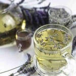 A glass cup filled with lavender tea, topped with lavender flowers, sits on a white surface. A teapot, another cup, and lavender sprigs surround it.
