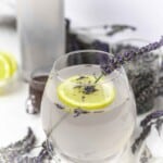 A glass of lemonade garnished with a lemon slice and a sprig of lavender, with more lavender and a jar of lavender in the background.