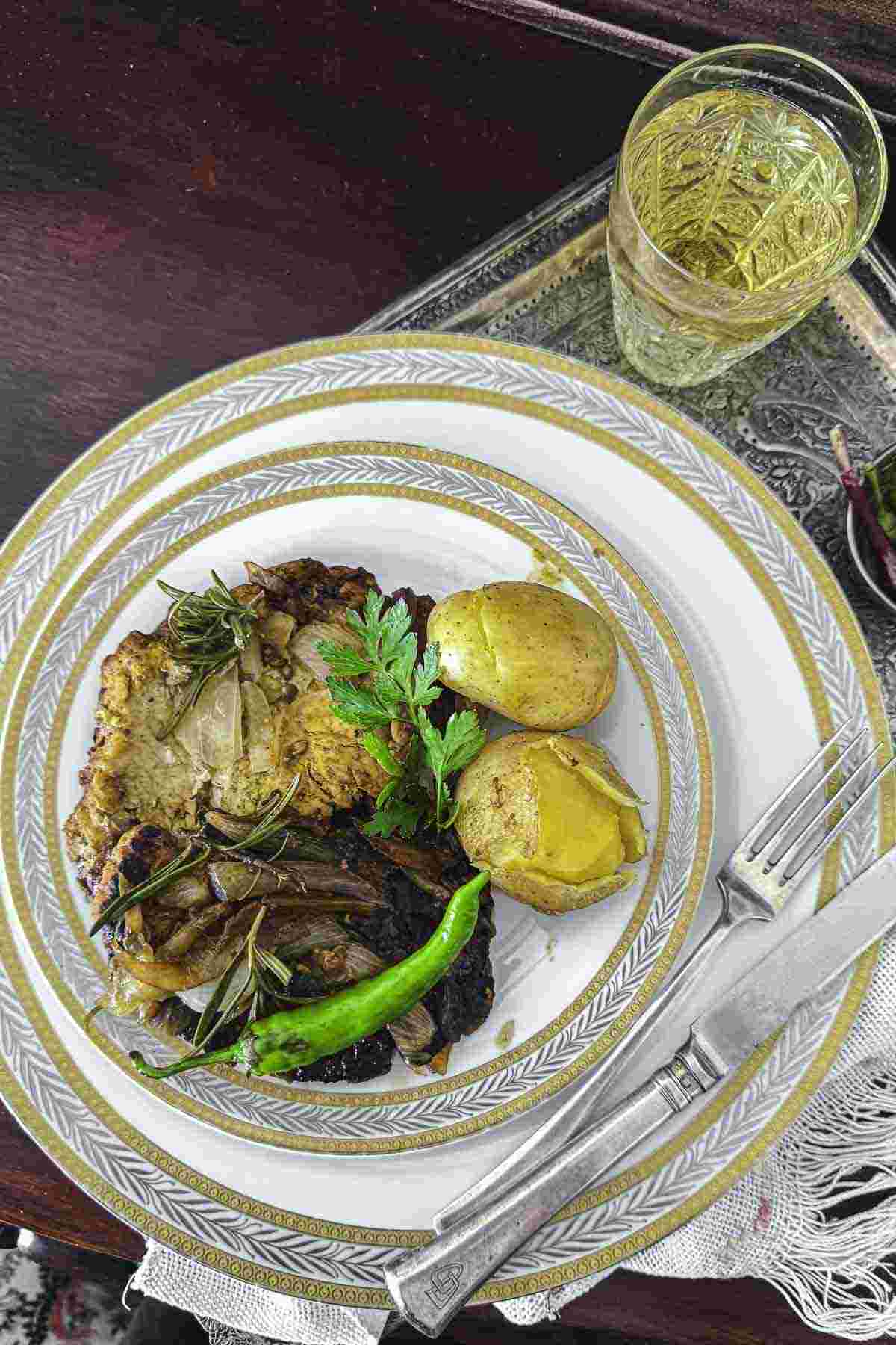 A plate with a pork fillet, roasted potatoes, and a green chili pepper, garnished with herbs. A clear glass with a yellow beverage is placed next to the plate.