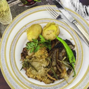 A plate with roasted meat, a baked potato, green chili, herbs, and sautéed onions. A fork and knife are placed beside the plate.