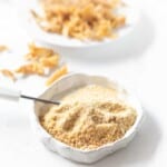 A small bowl filled with powdered and shredded dried chicken sits on a white surface. A knife is partially inserted into the powdered chicken. Another bowl with dried chicken pieces is in the background.