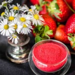 A small vase with white daisies stands next to a bowl of powdered strawberry, surrounded by fresh strawberries.