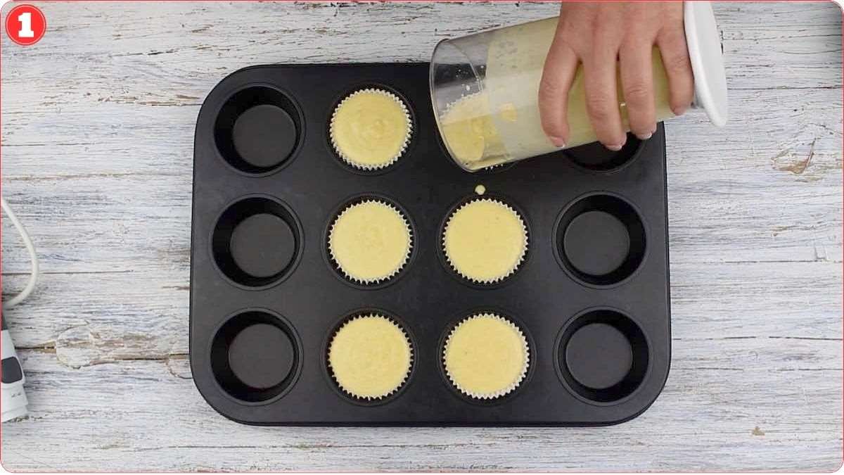 A person pours batter into paper liners inside a muffin tray, filling six of the twelve slots on a light-colored surface.