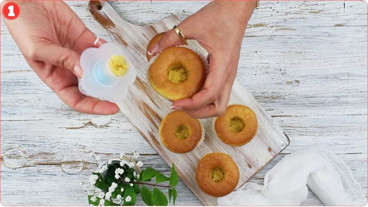 Hands are using a tool to fill the middle of cupcakes placed on a wooden board, with a small plant next to the board.