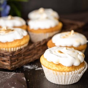 Several frosted cupcakes are placed on a dark surface. Some are arranged in a wicker basket while others are outside of it. The background is slightly blurred.