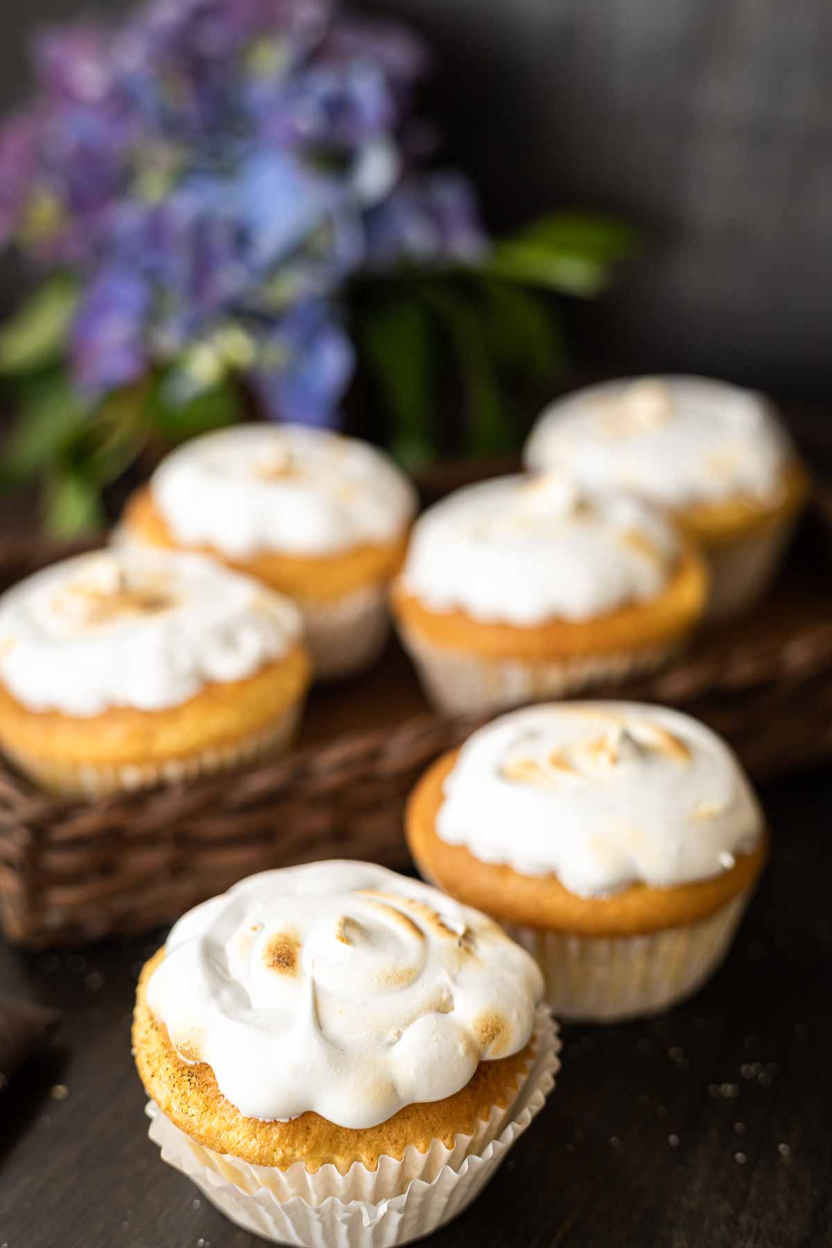 Six cupcakes with white frosting are arranged on a dark surface, with some of them presented on a woven tray. A blurred background features purple and green foliage.