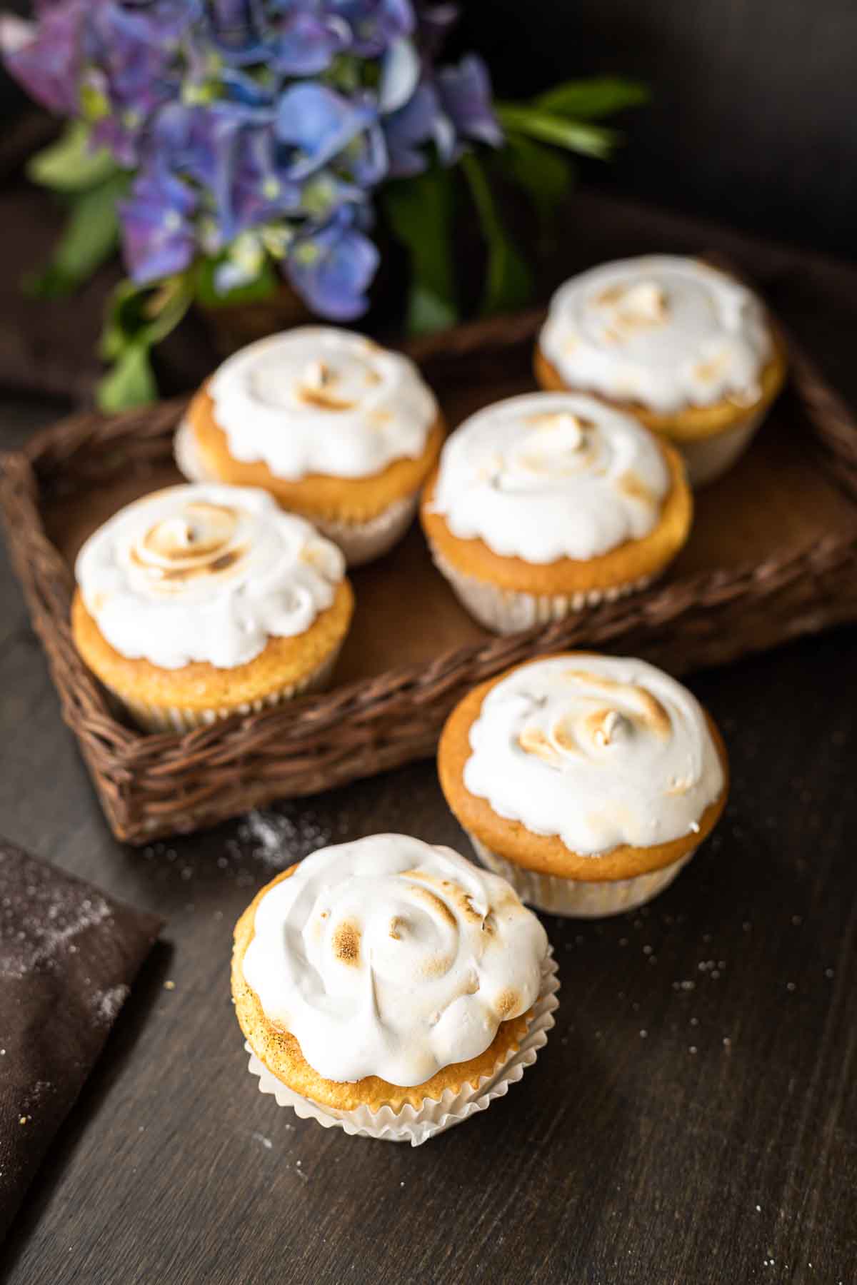 Six lemon meringue cupcakes with toasted tops are arranged on a dark wooden surface, some in a basket and others in front. A bouquet of purple flowers is in the background.