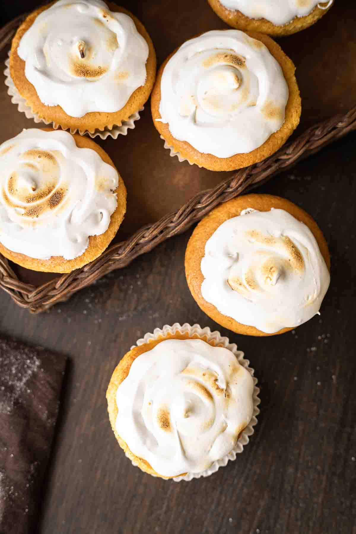 A basket and table hold six cupcakes topped with a white swirled frosting.