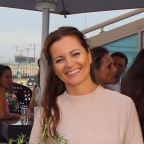 A woman with long brown hair smiles at a rooftop gathering, with people and cityscape visible in the background.