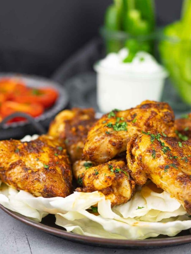 A plate of seasoned, roasted chicken wings garnished with herbs, served on a bed of cabbage, with a bowl of dip and fresh vegetables in the background.
