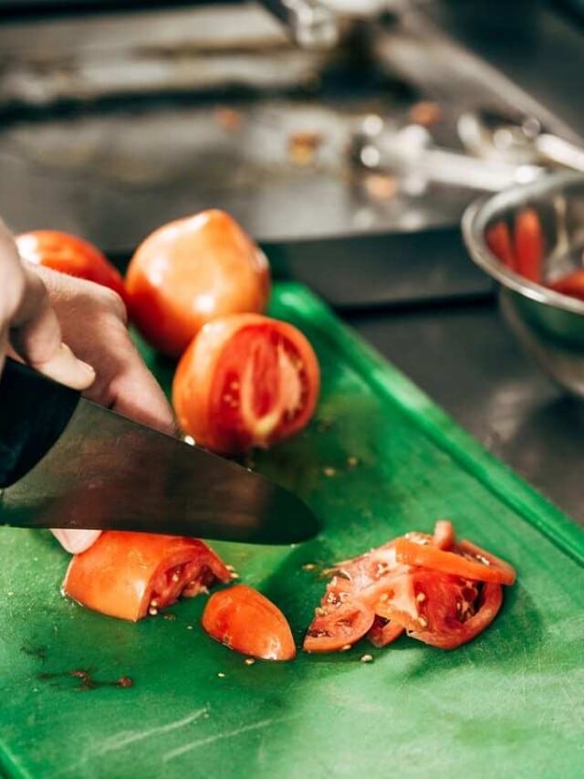 A hand using a knife to slice tomatoes on a green cutting board, with a metal bowl containing more tomato slices in the background.