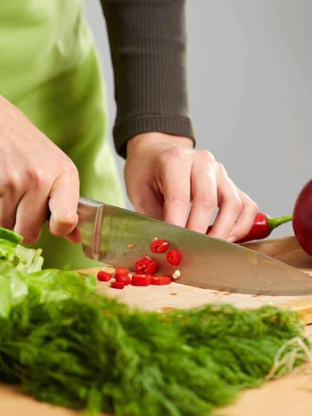 Person chopping red chili peppers on a wooden cutting board surrounded by fresh green herbs.