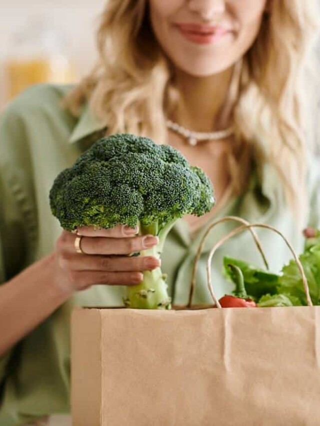 A person in a green shirt holds a piece of broccoli over an open paper grocery bag filled with various vegetables.