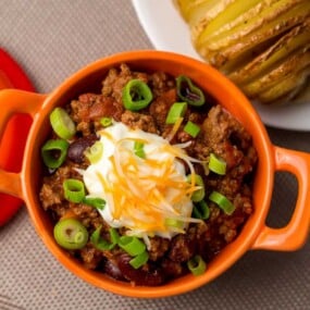 An orange bowl of chili topped with sour cream, shredded cheese, and green onions.