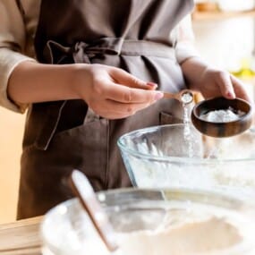 Measuring baking powder into a glass mixing bowl filled with ingredients.