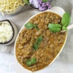 A dish of beef bolognese gravy garnished with basil leaves, served alongside bowls of shredded cheese and uncooked pasta, with purple flowers in the background.