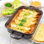 Baked enchiladas topped with melted cheese and herbs in a black baking dish. Accompanied by sliced avocado, lime wedges, and a small bowl of sauce.