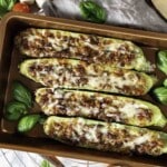Four stuffed zucchini halves in a baking dish, topped with melted cheese and herbs, surrounded by fresh basil leaves.
