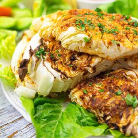 Grilled cabbage wedges garnished with herbs on a bed of lettuce, with sliced cucumbers and tomatoes in the background.