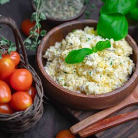 Two wooden bowls of creamy risotto garnished with basil leaves are next to a basket of cherry tomatoes and fresh herbs. Two wooden spoons are placed beside the bowls.