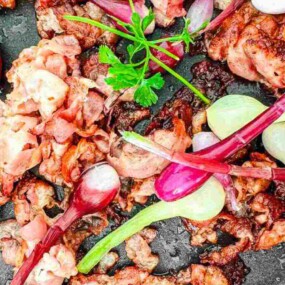 Fried bacon pieces and red onions with green stems on a black surface, garnished with a sprig of parsley.