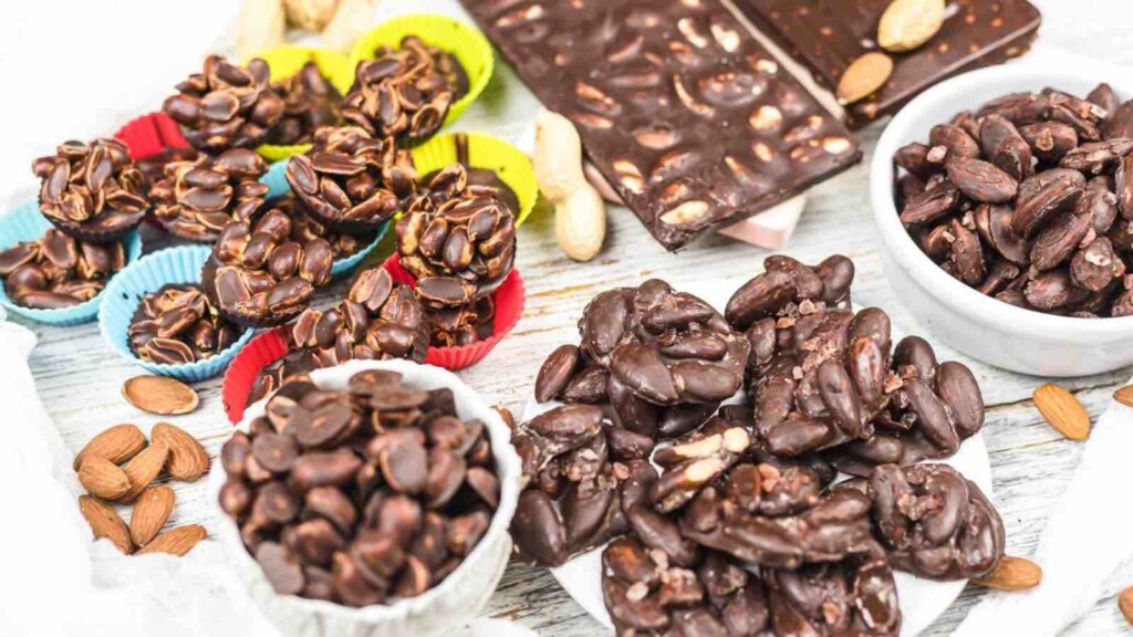 Various chocolate-covered nuts and clusters are displayed on a white wooden surface, including almonds and peanuts in colorful paper cups and white bowls.