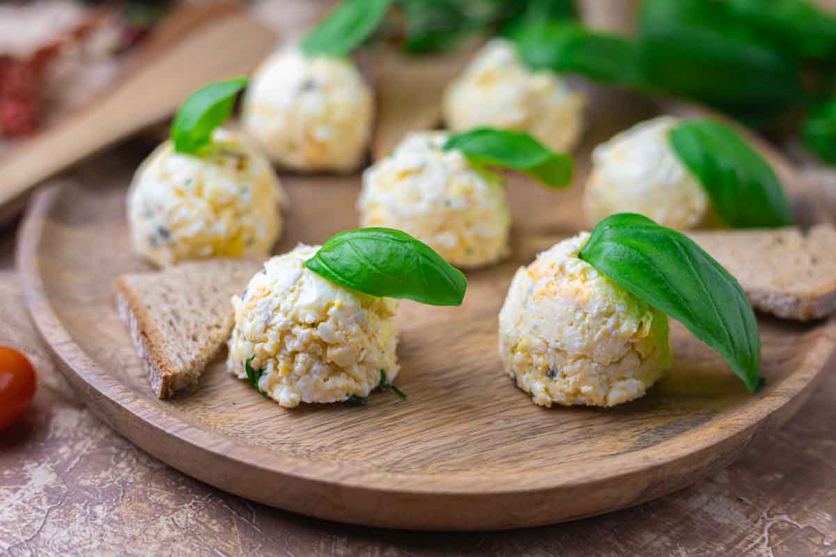 Cheese balls garnished with basil leaves on a wooden plate, accompanied by triangular pieces of brown bread.