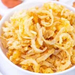 A bowl of crispy fried onions on a white plate with whole onions in the background.