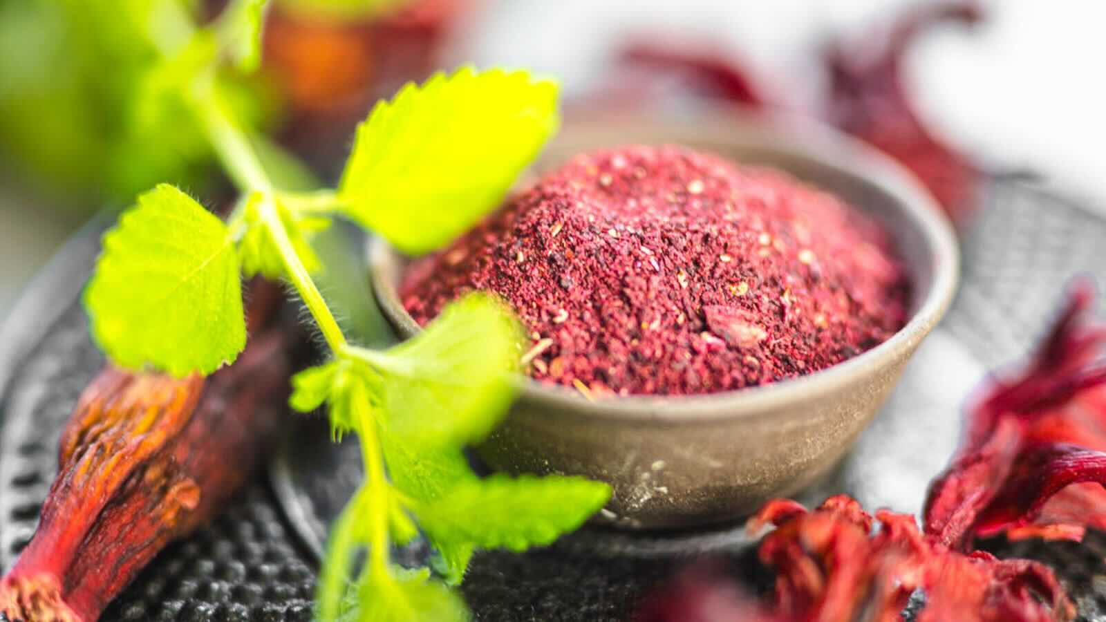 A bowl of ground hibiscus powder is surrounded by dried red hibiscus flowers.
