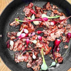 A frying pan on a wooden surface contains cooked bacon and small red onions.