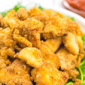 Close-up of a bowl of golden brown fried chicken pieces on a bed of greens, with a small bowl of red dipping sauce in the background.