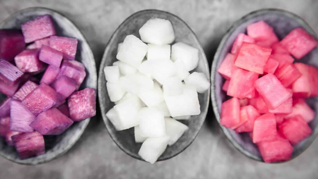 Three bowls of diced fruit or vegetables: purple cubes on the left, white cubes in the center, and pink cubes on the right, placed on a gray surface.