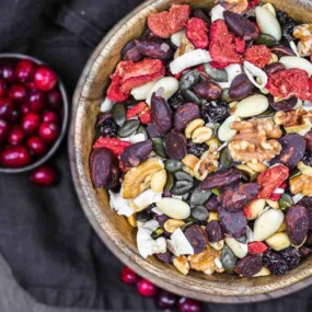 A wooden bowl filled with a mix of nuts, seeds, and dried fruits, placed on a dark cloth. A small bowl of fresh cranberries is beside it.