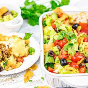 A casserole dish brimming with taco salad is topped with tomatoes, olives, and lettuce. Perfect for busy weeknights, an easy serving awaits on a separate plate with a fork. Fresh cilantro graces the background, adding a hint of vibrant green.