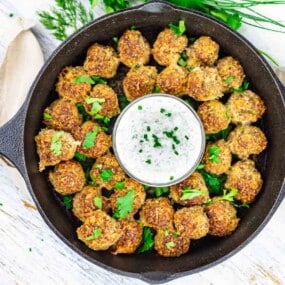 A cast iron skillet filled with browned meatballs arranged around a small bowl of creamy dip, garnished with chopped herbs.