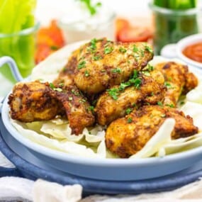 A blue plate with seasoned grilled chicken pieces on a bed of cabbage, garnished with herbs. Bowls of sauce and vegetables are in the background.
