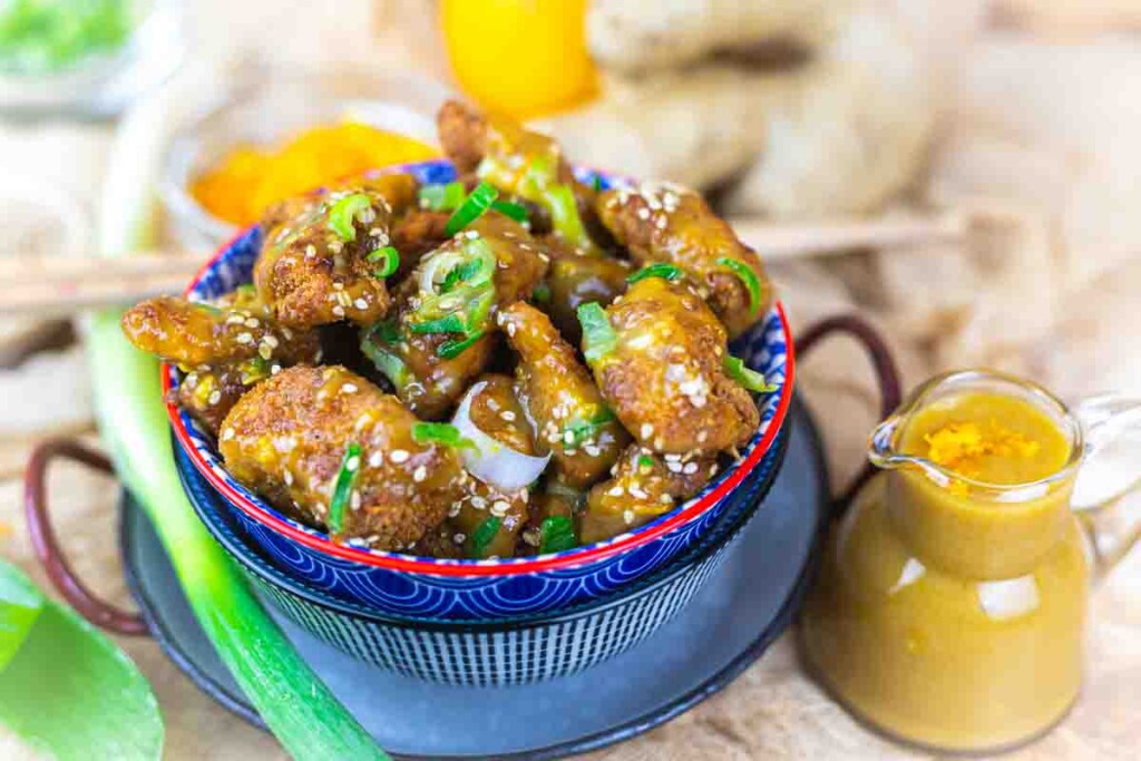 A bowl of fried chicken pieces topped with sesame seeds and green onions. A small pitcher of sauce is nearby, with a partially visible scallion and bell pepper in the background.