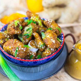 A bowl of fried chicken pieces topped with sesame seeds and green onions. A small pitcher of sauce is nearby, with a partially visible scallion and bell pepper in the background.