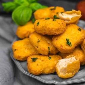 A plate of golden-brown chicken nuggets garnished with herbs. Some nuggets are broken open, revealing the white chicken inside, with green leaves in the background.