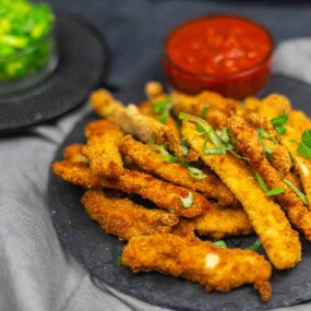 A plate of breaded, fried zucchini sticks garnished with herbs, accompanied by a bowl of red sauce and a bowl of chopped green vegetables in the background.