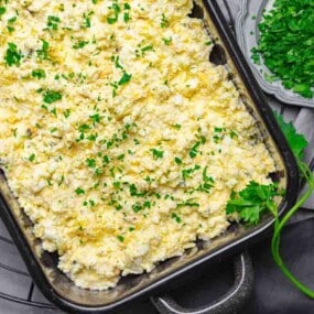 A baking dish filled with mashed potatoes topped with chopped parsley. Nearby, a bowl of more parsley and a few cherry tomatoes and cilantro leaves are placed on a gray cloth.