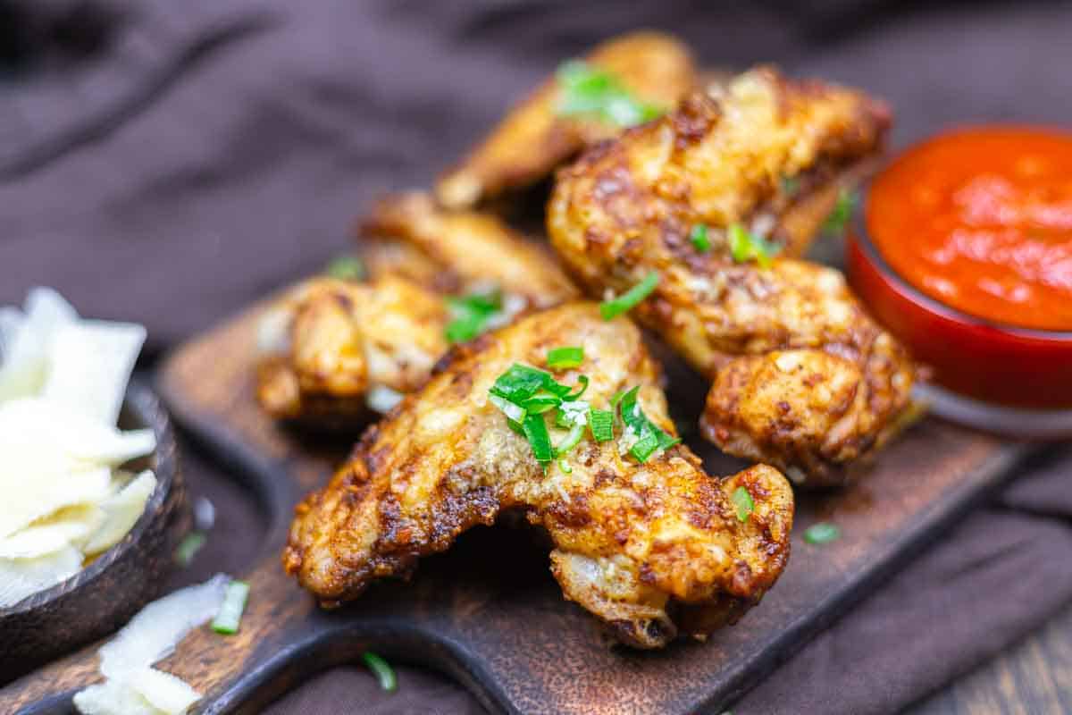 Four seasoned chicken wings garnished with chopped herbs on a wooden board, served with a side of red sauce and shredded cheese.