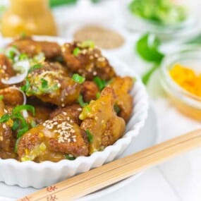 A bowl of sesame-covered, glazed fried chicken pieces garnished with chopped green onions. Chopsticks rest on the bowl's edge. Ingredients like sesame seeds and peppers are visible in the background.
