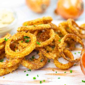 A pile of crispy onion rings garnished with chives, served with small bowls of dipping sauce and ketchup on the side.
